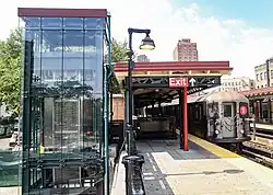 Panorama einer Station bei Tageslicht. Links ein neuer, vollverglaster Personenaufzug. Mittig ein beige und rot gestrichenes Bahnsteigdach aus Holz im Craftsman-Stil. Rechts die Gleise, darauf ein haltender Zug der Linie 1. Im Hintergrund links Bäume, rechts mehrgeschossige Altbauten und dunkelrot backsteinverschalte Punkthochhäuser.
