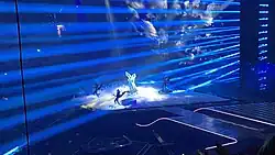 Foto von einer Bühne mit einer in einem weißen Engelskostüm gekleideten Person im Zentrum, die von schwarz gekleideten Teufeln umgeben ist