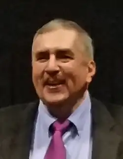 Mann mittleren Alters von vorn im dunklen Jackett, hellem Hemd und rot-pinker Krawatte in die Kamera schauend in Farbe.