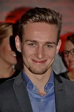 Floors schaut lächelnd in die Kamera. Er hat kurzes blondes Haar und trägt ein aufgeknöpftes hellblaues Hemd und ein graues Jackett.