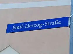 blaues, rechteckiges Straßenschild mit der Aufschrift „Emil-Herzog-Straße“ vor einer Hauswand