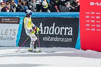 Das Bild zeigt Skifahrerin Emma Aicher in Andorra bei einem Skirennen Grandvalira 2023.Emma Aicher im Vordergrund. Sie trägt einen gelben und weißen Rennanzug, einen Helm und Skibrillen. Ihre Skier sind mit dem Markennamen "HEAD" versehen. Sie hält zwei Skistöcke mit rosa Griffen. Im Hintergrund sieht man eine Menge Zuschauer, die das Rennen beobachten. Hinter den Zuschauern ist eine große, blaue Werbewand mit dem Namen "Andorra" und der Webseite "visitandorra.com" zu sehen. Ein Teil einer weiteren roten Werbewand mit dem Audi-Logo ist rechts im Bild zu sehen. Der Schnee auf dem Hang ist frisch und weiß, und die allgemeine Atmosphäre ist die eines professionellen Skirennenveranstaltung bei hellem Tageslicht.