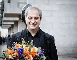 Brustbild eines in die Kamera lächelnder Mannes mit grauen Haaren und schwarzem Hemd und Anzug. Der auf dem Rücken getragene schwarze Cellokoffer schaut hinter dem Kopf hervor. Er hält einen bunten Blumenstrauß in der Hand. Im Hintergrund die Rückseite des Kölner Rathauses