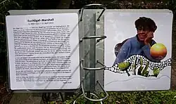 Die Tafel ist aus silberfarbenen Metall und zeigt eine Spiralbindung, die zwei aufgeschlagene laminierte Seiten zeigt. Die linke Seite enthält einen Text, die rechte Seite zeigt ein Porträt von Ika, das von verschiedenen Elementen, wie einer Kugel und Linien abstrakt gestaltet ist.