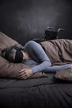 Eine an ME/CFS erkrankte Person liegt auf der Seite in einem Bett. Sie ist mit einer Decke zugedeckt. Die Person trägt einen Gehörschutz und eine Schlafmaske. Im Hintergrund steht ein Rollstuhl.