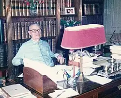 Ernest Labrousse lehnt sich vor einem Bücherregal und hinter einem hölzernen Schreibtisch in seinen Stuhl