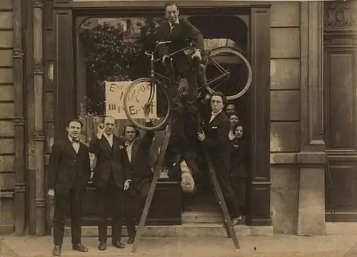 Freunde von Max Ernst vor der Ausstellung in der Pariser Galerie Au Sans Pareil, 1921. Auf der Leiter mit Fahrrad steht Philippe Soupault, kopfüber hängend Jacques Rigaut, rechts auf der Leiter André Breton.