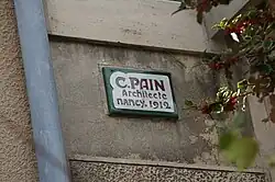 Namenszug des Architekten nebst Jahresangabe 1912 und der Ort Nancy auf einem weißen Hintergrund mit grüner Umrandung an einer Hauswand.