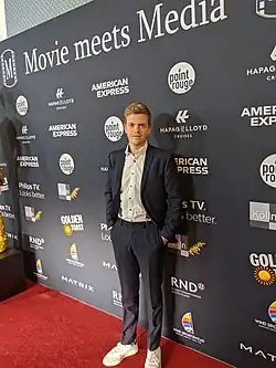 Ferdinand Hofer auf dem roten Teppich beim „Movie meets Media“-Event in Hamburg im Jahr 2019. Er trägt einen dunkelblauen Anzug mit einem weißen Hemd und weißen Sneakers. Der Hintergrund besteht aus einer Medienwand mit verschiedenen Sponsorenlogos, darunter „American Express“, „Point Rouge“, „Hapag-Lloyd Cruises“, „Golden Toast“, „Philips TV“ und weitere. Die Medienwand ist dunkel mit weißer Schrift, und der Eventname „Movie meets Media“ ist oben deutlich sichtbar. Ferdinand Hofer steht mit den Händen in den Taschen und posiert für ein Foto.