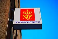 Foto eines an einem Gebäude angebrachten Schildes mit einem Logo mit rotem Hintergrund, dass zwei goldene Fische und eine Meerjungfrau unter einer goldenen Krone zeigt