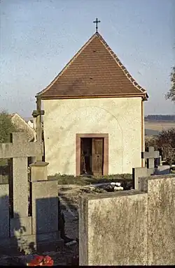 Über der Eingangstür sieht man einen Halbkreis, das war der frühere Anschluss an das Langhaus. Vor der Eingangstür steht ein steinernes Kreuz im Querprofil sichtbar. Das Kreuz bildete den Abschluss der Sichtachse von dem früheren Seiteneingang zum Friedhof. Der Verbleib diese Kreuzes ist ungeklärt.
