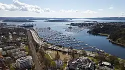 Foto eines Küstenbereichs. Direkt an der Küste verläuft eine Autobahn, nahezu parallel dazu verlaufen Schienen, dahinter befinden sich Wohnhäuser. Im Fjord befinden sich viele Anlegeplätze für Boote
