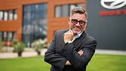 ein freudlos grinsender bärtiger Mann mit Brille in einem Anzug