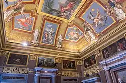 Farbige Deckenmalerei mit mehreren Szenenfeldern, auf denen antike Menschen und Himmelswesen agieren. Die Zwischenräume sind mit goldenen Ornamenten und Hieroglyphen ausgefüllt. Ägyptische Figuren werden am Deckenrand von Putten mit Blumenkränzen an die Felder verbunden und wirken dreidimensional. Die angrenzenden Wandseiten haben Ölgemälde und ägyptische Reliefs.