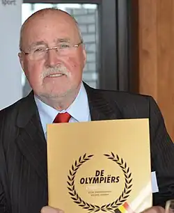 Farbfoto vom 75-jährigen Gaston Roelants im Anzug, in der Hand das Buch Olympiërs 1900-2012, das er als Vorsitzender des Vereins belgischer Olympiateilnehmer präsentiert.