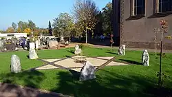 In die Erde eingelassene Eisenbahnschienen bilden das Symbol des Davidstern. Sechs Findlinge sind zu sehen, die an den Sternenden aufgestellt sind. Um diese Installation ist grünes Gras zu sehen. Im Hintergrund sind Grabsteine des Friedhofes erkennbar Am rechten oberen Bildrand ist ein Teil der Kirche St. Martin zu sehen.