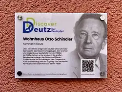 Gedenktafel an einer roten Wand mit der Überschrift „Wohnhaus Otto Schindler – Karneval in Deutz“. Die Tafel ist Teil des Projekts „Discover Deutz – Der Deutzpfad“. Auf der linken Seite steht ein Text, der erklärt, dass der Grafiker und Wagenbauer Otto Schindler über Jahrzehnte das Bild des Kölner Rosenmontagszugs mitgestaltete. Auf der rechten Seite ist ein Porträtfoto von Otto Schindler zu sehen. Unten rechts befindet sich ein QR-Code, links unten das Logo des Projekts und ein Hinweis auf die Bürgervereinigung Deutz als Initiator.
