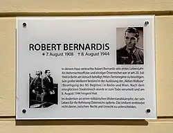 Gedenktafel angebracht an einer gelb bemalten Hausmauer mit zwei historischen Fotografien und Texten.