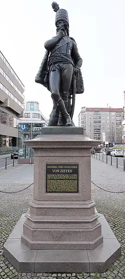 Farbige Untersicht auf eine Männerfigur mit Uniform, der seine Beine überkreuzt und sich mit der linken Hand am Dolch abstützt. Mit der rechten Hand reibt er an seinem Kinn und blickt nach links. Die Figur steht auf einem Sockel mit der Aufschrift „General der Cavallerie von Zieten“.