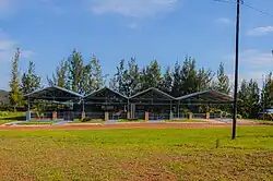 Blick über eine dünn mit kurzem Gras bewachsene Fläche frontal auf die Gedenkstätte. Diese ist eingezäunt und mit einem metallenen Paralleldach ausgestattet. Rechts neben der Gedenkstätte steht ein Flaggenpfosten mit der ruandischen Flagge.