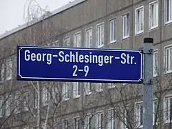 Blaues Straßenschild mit weißer Schrift. Aufschrift: Georg-Schlesinger-Str. 2–9. Im Hintergrund sind kahle Bäume vor einem Plattenbau zu sehen