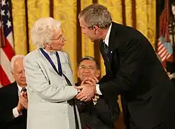 Im Vordergrund stehen links Ruth Johnson Colvin und rechts der damalige US-Präsident George W. Bush. Colvin trägt ein hellgraues Kleid mit farblich passendem Jackett und das blaue Band der Presidential Medal of Freedom um den Hals. Sie ist weißhaarig und hat ihren Kopf leicht zu Bush gedreht. Der grauhaarige Bush trägt einen dunklen Anzug mit weißem Hemd und blauer Krawatte. Beide sprechen miteinander. Bush steht leicht gebückt und hält Colvins Hände mit seinen Händen umfasst. Hinter ihnen sitzen David McCullough und Norman Mineta, zwei weitere Empfänger der Medaille, die ausschnittsweise zu sehen sind. Sie tragen Anzüge und applaudieren. Den Hintergrund bilden ein geschlossener gelber Fenstervorhang und am linken Bildrand eine amerikanische Flagge.