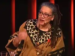 Bild des Oberkörpers einer Frau, die in eine Headsetmikrophon spricht und sich leicht zur Seite neigt. Sie trägt afrikanisch anmutende Kleidung und Schmuck und trägt ihr Haar in nach hinten gebundenen Rastalocken.