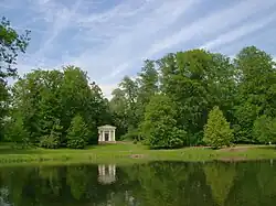 Frontale Farbfotografie eines kleinen, hellen Säulentempels, das zwischen den Bäumen leicht verborgen ist. Die Bäume nehmen die ganze mittlere Bildebene ein. Im Vordergrund ist eine Wasserfläche.