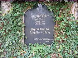 Schwarzer Grabstein mit goldener Schrift, an einer von Efeu bewachsenen Mauer angebracht. Die Inschrift lautet: Auguste Löber, geb. Feige, geb. 22. Nov. 1824, gest. 15. Jan. 1897, Begründerin der Auguste-Stiftung, „Selig sind die Bamherzigen, denn sie werden Bamherzigkeit erlangen.“, Matth. 5,7