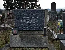 Schwarzer Grabstein mit weißer Musterung von vorn mit Sterbedaten von drei weiteren Personen auf einem Sockel.