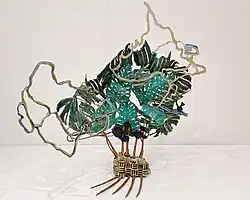 Bronzeskulptur mit üppigen grünen Blättern einer Monstera-Pflanze und mäandrierenden kartografischen Linien, sowie perforierte blauen Abstraktionen aus Karten des Meeresbodens.