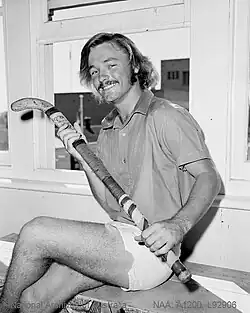 Greg Browning sitzt auf diesem Schwarzweißfoto auf einer Holzbank vor einem Fenster. Er trägt eine weiße Hose, ein farbiges Trikot und in seinen Händen einen Hockeyschläger.