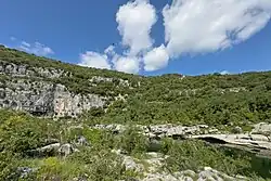 Blick vom linken Ufer der Ardèche zur Grotte Chabot. Der Eingang ist durch Büsche und Bäume verdeckt