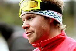 Seitliches Porträt von Tomas Gustafson. Seine gelbe Skibrille sitzt auf der Stirn, darunter ein buntes Stirnband. Außerdem ist der Kragen einer roten Jacke oder eines roten Anzugs zu sehen.