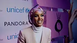Halima Aden trägt ein rosafarbenes Kopftuch und cremefarbene Kleidung. Im Hintergrund ist eine Wand mit Werbung zu sehen.