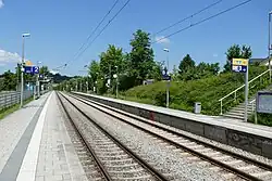 In der Bildmitte verlaufen zwei parallele Gleise, links und rechts davon jeweils ein typischer S-Bahnsteig mit Schildern, Uhr, Mülleimer etc. Hinter den Bahnsteigen sieht man jeweils eine begrünte Umgebung.