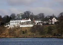 Südost-Ansicht des Hotelrestaurants Jacob von der Elbe aus. Am Ufer bildet der Elbuferweg einen kurzen Abschnitt des Elbuferwanderweges, der hier vorbeiführt. Rechts ist die Jacobs Treppe zu erkennen, die hier von der Höhe der Elbchaussee hinunter zum Ufer führt. Bei dieser Treppe erinnert ein Gedenkstein an die Primus.