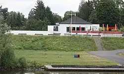 Im Foto ist vorn der Main, dann der Rudersteg und dahinter ein Deich. Vom Bootshaus hinter dem Deich ist nur die Gastronomie im ersten Stock erkennbar, darunter sind die Bootshallen mehr zu erahnen.