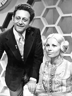 Hans Rosenthal und Monika Sundermann in der Sendung Dalli-Dalli (1971) Hans Rosenthal trägt einen dunklen Anzug mit einer gemusterten Krawatte. Er hat dunkles, welliges Haar und steht leicht nach vorne gebeugt. Monika Sundermann auf der rechten Seite sitzt und trägt ein Kleid mit einem komplexen Muster sowie eine Perlenkette. Ihr Haar ist hell und zu einem stilvollen Knoten hochgesteckt. Der Hintergrund besteht aus einer Wand mit sechseckigen Muster. Diese Muster waren typisch für die Sendung Dalli Dalli.