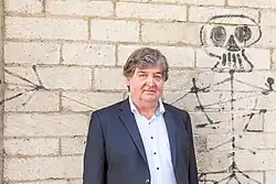 Ein Mann im Anzug und mit gewelltem Haar steht vor einer mit Werksteinen gemauerten Wand, auf die ein schwarzes, leicht verblasstes Skelettgraffito gesprayt ist. Es sieht aus, als ob das Skelett den rechten Arm um den Mann legt.