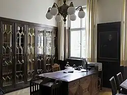 Schwere dunkle Möbel: Ein Schreibtisch und mehrere Aktenschränke mit Glasfronten, an der Wand ein Porträt von Prinzregent Luitpold. Das Büro wird weiterhin genutzt, deshalb stehen auf dem alten Schreibtisch ein Computer und ein Telefon, an der Wand ein Drucker