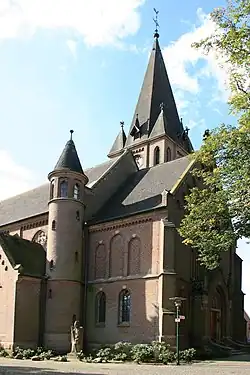 Das Bild zeigt eine Kirche aus Ziegelsteinen mit schwarzen Dachpfannen. Vor ihr steht eine kleine Statue.