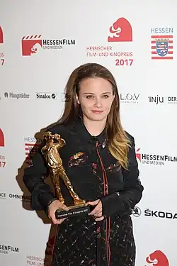 Jasna Fritzi Bauer mit langen, braunen Haaren und trägt ein schwarzes Hemd mit bunten Details. Im Hintergrund sind Logos und Schriftzüge des Hessischen Film- und Kinopreises 2017 zu sehen. Sie hält eine goldene Trophäe in Form einer stehenden Figur mit einem Hut in der Hand. Die Plakette auf der Trophäe trägt die Aufschrift „Hessischer Film & Kinopreis 2017“. Im Hintergrund sind verschiedene Logos und Schriftzüge zu sehen, darunter „HessenFilm und Medien“, „Hessischer Film- und Kinopreis 2017“, „Hessen“, „L.O.V Cosmetics“, „Inju“, „Der Medien“, „Pfungstädter“, „Sinalco“, „Skoda“ und „Omnimago“.