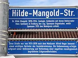 Foto des Straßenschilds in der Hilde-Mangold-Straße mit Erörterung darunter: Diese Straße war von 1931 bis 2018 nach dem Mediziner Alfred Hegar benannt – einem wichtigen Vertreter des Sozialdarwinismus. Die Umbenennung erfolgte aufgrund seiner Befürwortung und Verbreitung einer negativen rassehygienisch begründeten Erbgesundheitslehre.
