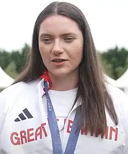 Holly Dunford trägt eine weiße Trainingsjacke mit der Aufschrift Great Britain. Um den Hals hängt ein blaues Band, an dem ihre olympische Medaille befestigt ist, die allerdings nicht im Bild zu sehen ist.