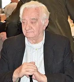 Miklòs Holop sitzt bei einem Empfang. Er trägt ein dunkles Jacket und ein helles Hemd. Seine Hände stützt er auf einen Gehstock.