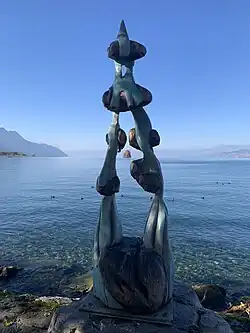 Blick auf den Genfersee bei Sonnenschein im Herbst durch eine Holzskulptur in Braun- und Grüntönen. Im Hintergrund ist die Ile de Peilz zu erkennen.