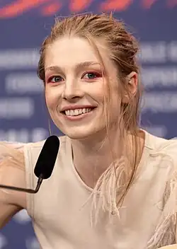 Hunter Schafer spricht lächelnd auf der Bühne einer Pressekonferenz der Berlinale 2024 in ein Tischmikrofon. Ihre blonden Haare sind nach hinten gebunden, einzelne Strähnen fallen seitlich an ihren Wangen vorbei.
