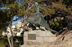 Bronzestatue, die einen auf einem Delphin reitenden Jungen zeigt. Im Hintergrund Kiefern und der Hafenort Hydra.
