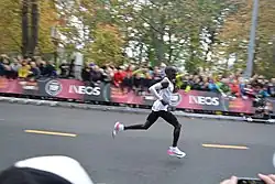Eliud Kipchoge läuft von links nach rechts durch das Bild. Kipchoge trägt ein weißes Oberteil, weiße Armstulpen, eine enge schwarze Hose und weiße Schuhe mit einer auffälligen pinken Applikation. Im verschwommenen Hintergrund sind Menschen zu sehen, die Kipchoge vom Streckenrand anfeuern.